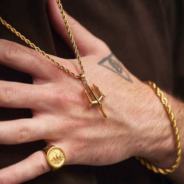 Hand wearing a gold necklace with a trident pendant and a gold ring on a dark background