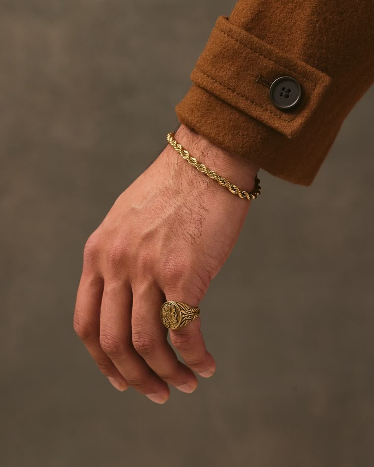 Hand wearing a gold ring and bracelet with a brown coat sleeve on a neutral background